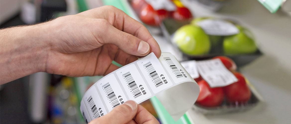 Main décollant une étiquette de prix avec code-barres dans une épicerie avec des pommes et des tomates en arrière-plan