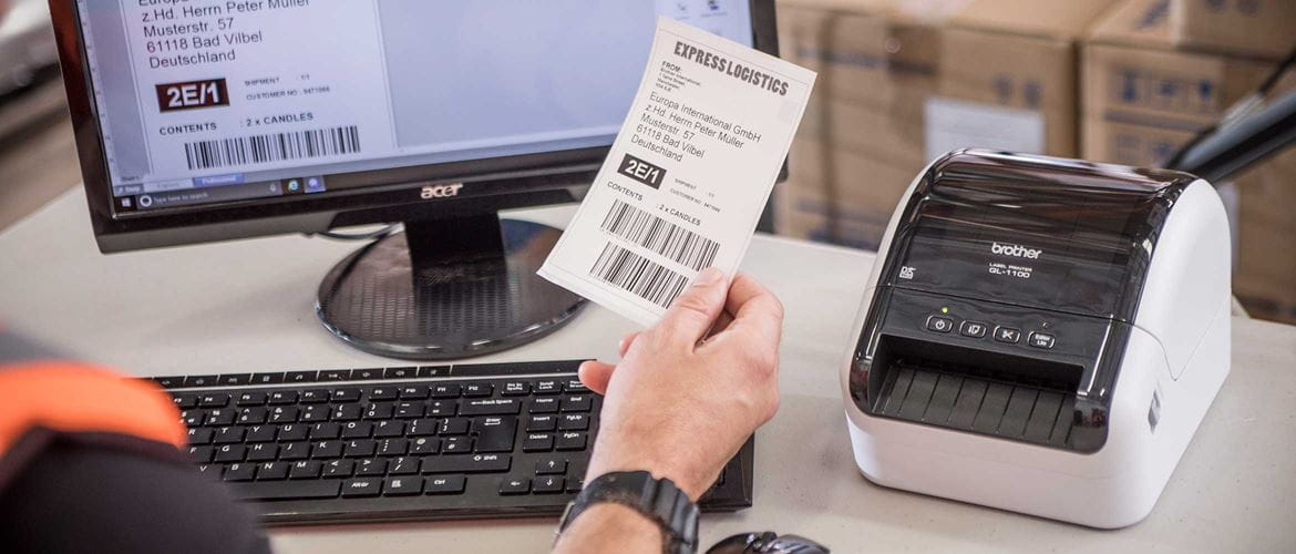 De Brother QL-1100 desktop labelprinter met een persoon die aan het bureau zit en het etiket vasthoudt