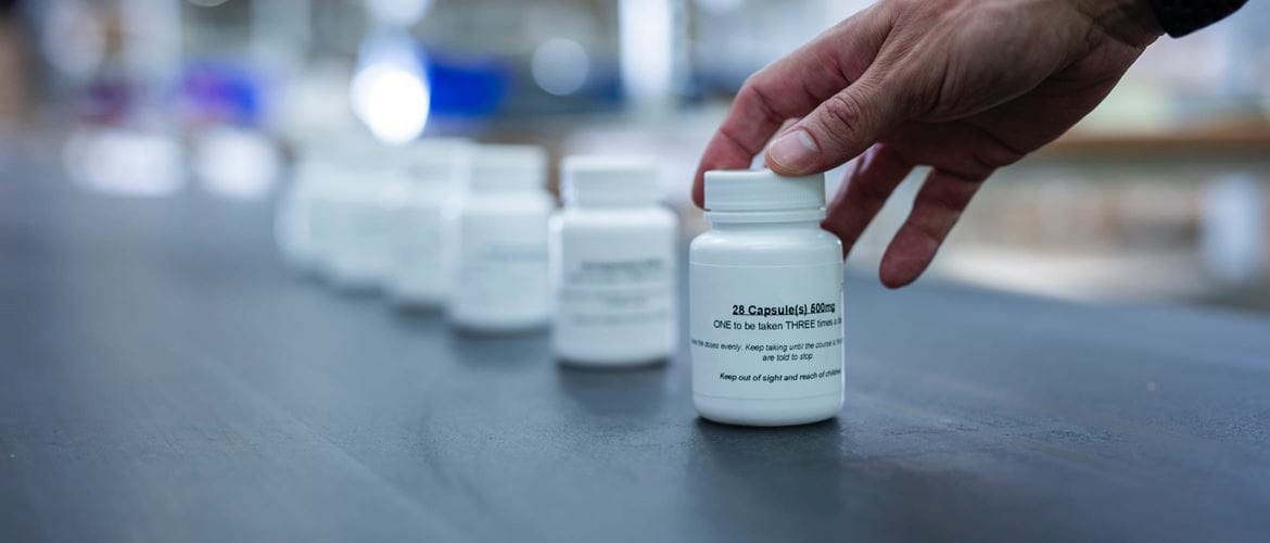 A factory worker picking up a white labelled pharmaceutical capsule container from a production line