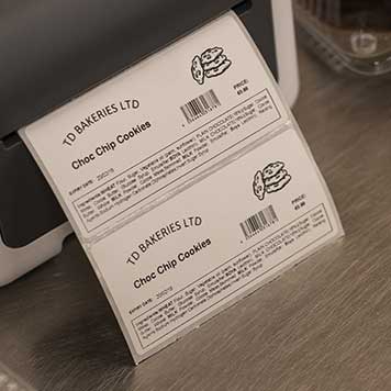 Grey and white Brother printer on stainless steel bench with printed label, chococlate muffin, clear plastic box