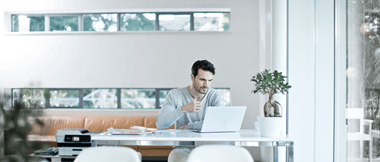 Een man zit aan zijn bureau in een thuiskantoor in een lichte kamer met twee planten. Een kleine printer staat op het bureau.
