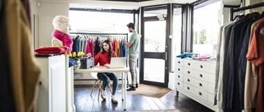 Een vrouwelijke medewerkster van een kledingwinkel zit aan een bureau om een document uit een Brother-printer te halen, terwijl een mannelijke shopper achter haar aan een kledingrek staat.