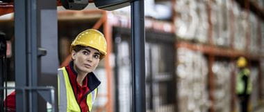Une employée d'une usine ou d'un entrepôt porte un casque de sécurité et une veste haute visibilité, tandis que des stocks sont visibles à l'arrière-plan de cette scène de transport et de logistique