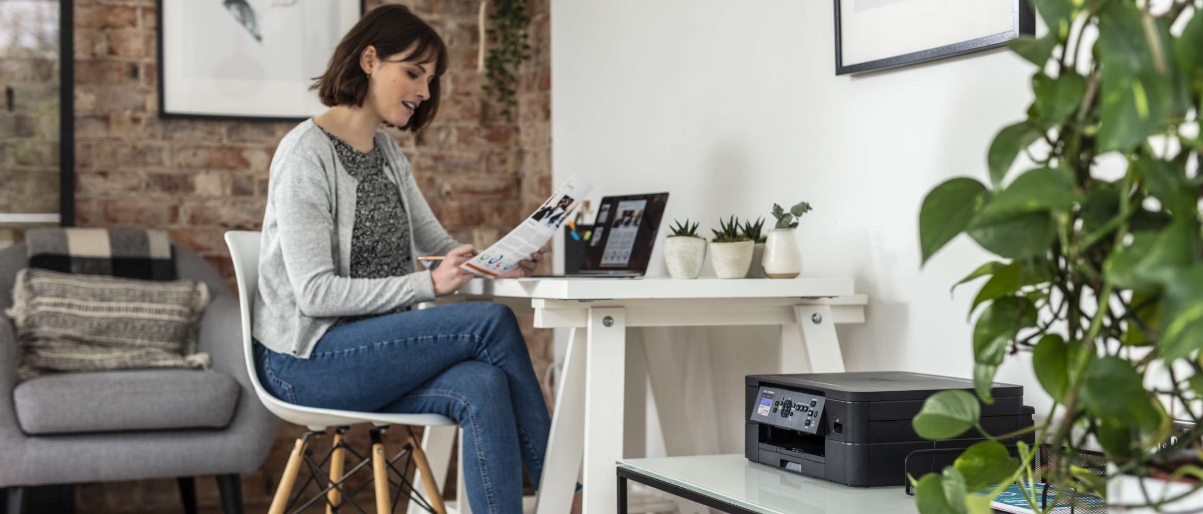 Een vrouw zit in haar thuiskantoor, van thuis uit te werken op haar laptop. Ze bekijkt een bedrijfsdocument dat ze heeft afgedrukt met haar compacte Brother-printer die op de voorgrond staat. 