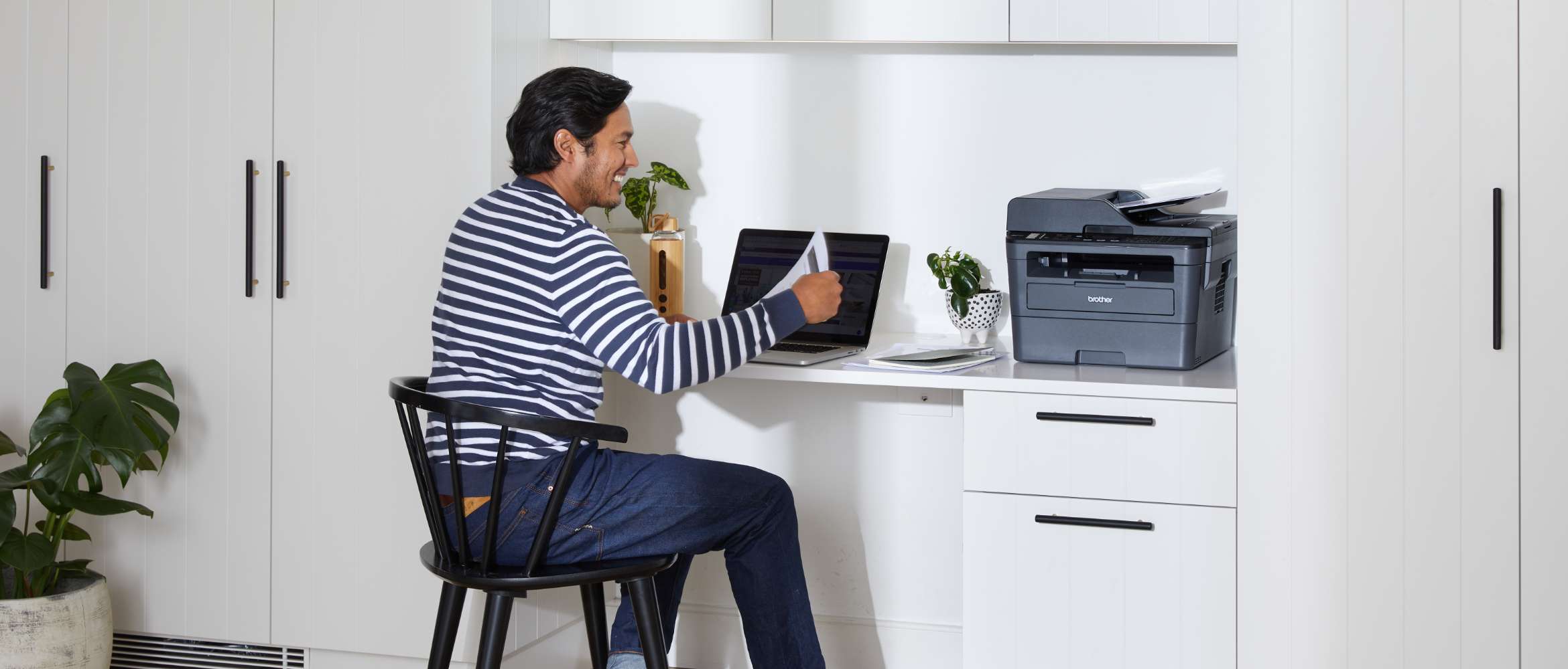 Un homme est assis dans son bureau hybride où il travaille à distance depuis son domicile. Il examine un document imprimé à partir d'une imprimante Brother.