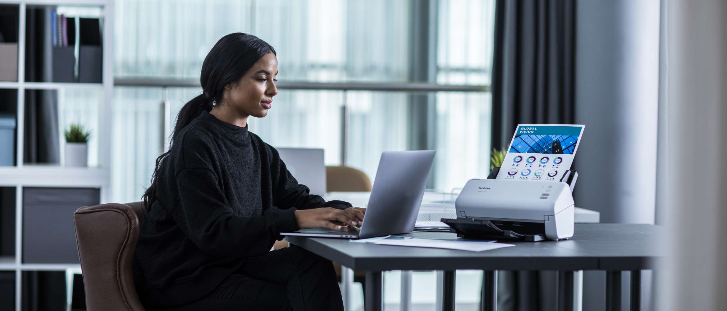 Une employée du secteur juridique est assise à un bureau et travaille sur son ordinateur portable, avec un scanner de documents Brother ADS à ses côtés.