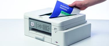 Een hand neemt een heldere indigo, groene en witte afdruk van de bovenkant van een witte Brother printer