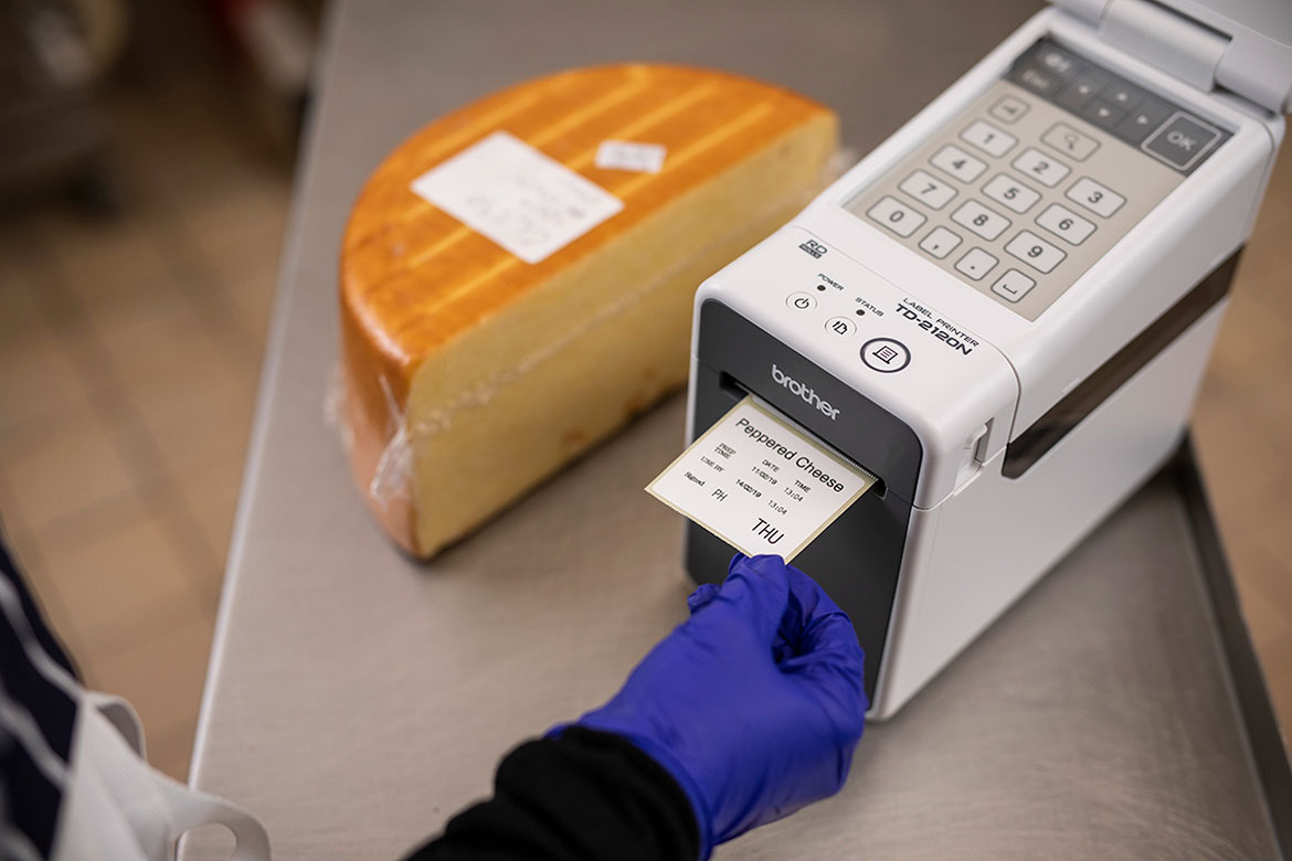 Hand in hanschoen neemt voedseletiket uit Brother TD printer in een keuken
