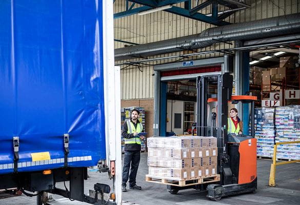 Woman driving fork lift truck with pallet into a truck in warehouse and distribution centre loading bay, man stood with hi-vis holding clipboard
