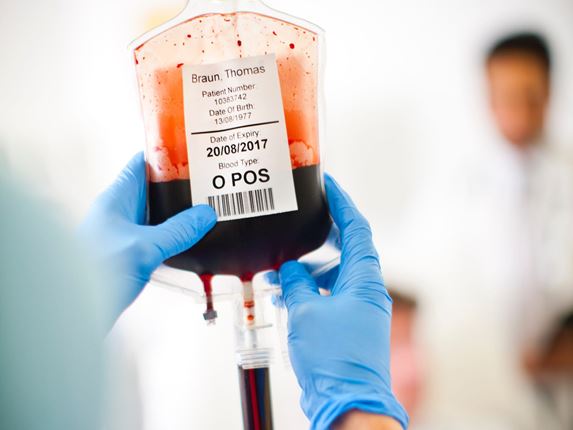 Une infirmière portant des gants chirurgicaux bleus lit les informations relatives à un patient sur une étiquette imprimée sur une poche de sang, avec un médecin en arrière-plan.
