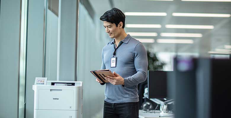Man wearing lanyard holding tablet next to Brother printer in office