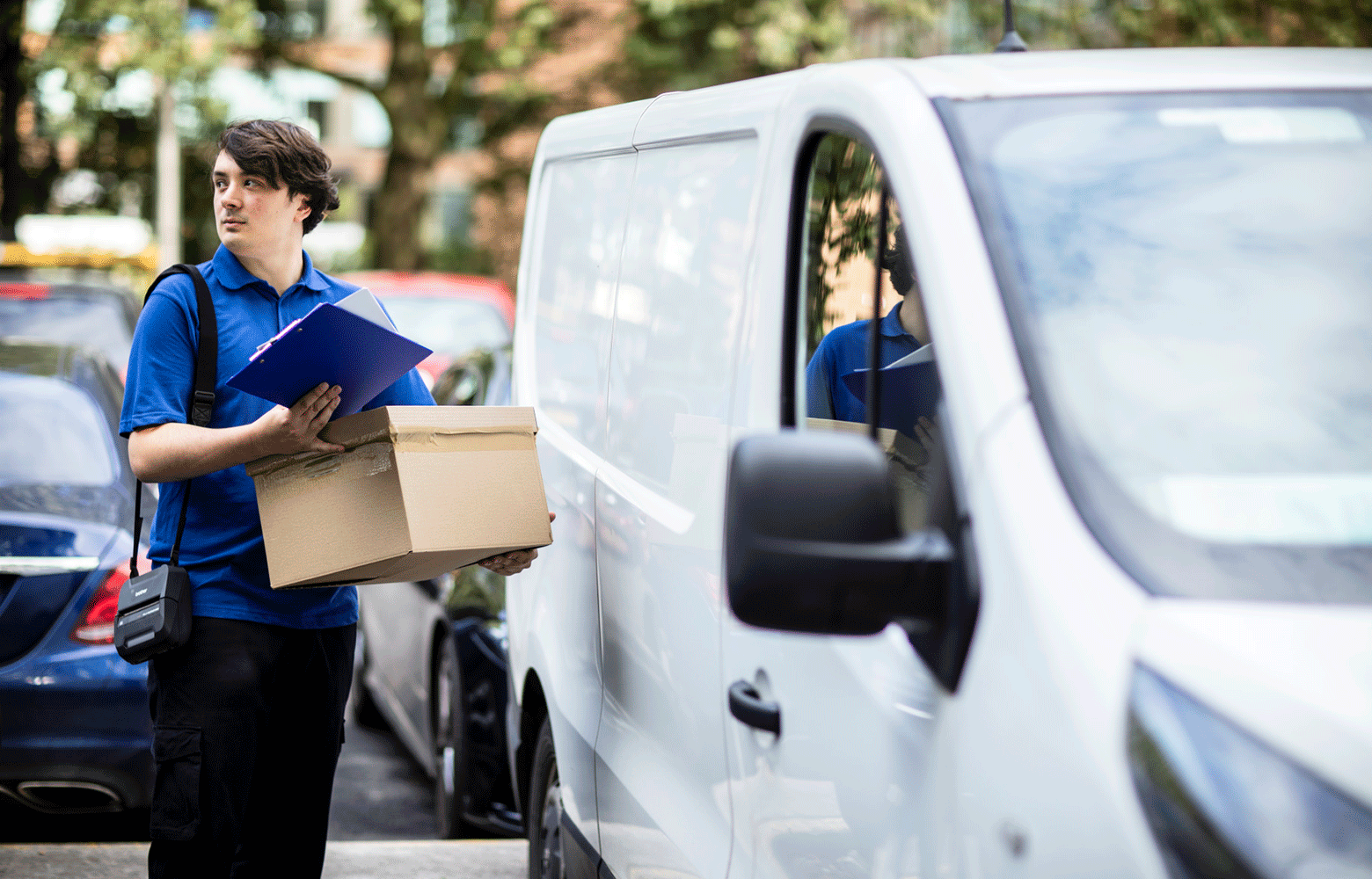 Un livreur tenant une boîte et un presse-papiers avec une imprimante RJ en bandoulière près d'une camionnette blanche