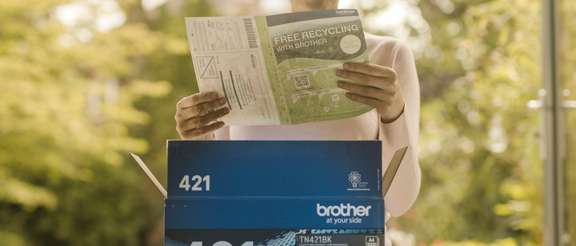 A woman reading recycling instructions on a green paper behind a blue, Brother toner cartridge box