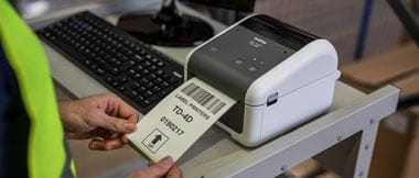 A Brother label printer on a desk using thermal printing technology to print a medium sized white label with black text.