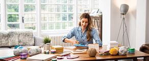 Kobieta korzysta z nożyczek siedząc przy stole, na którym stoją słoiki z dżemami, pudełka, motki wełny i akta osobowe 