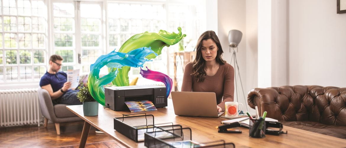 Imprimanta din care ies valuri viu colorate de cerneală în timp ce imprimă lângă un bărbat și o femeie care lucrează de acasă