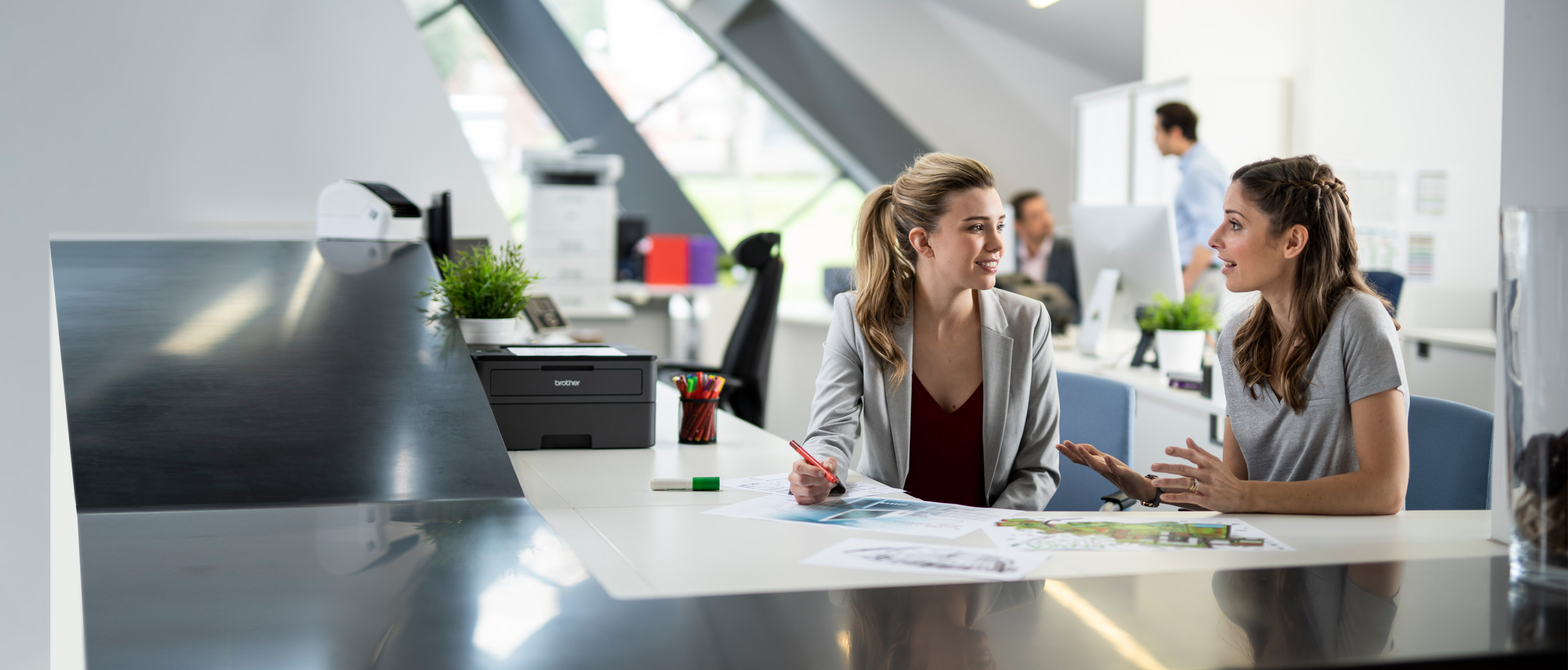 Two woman sat in a busy office at a desk talking, men in the background, printers, pens, pen pot, documents