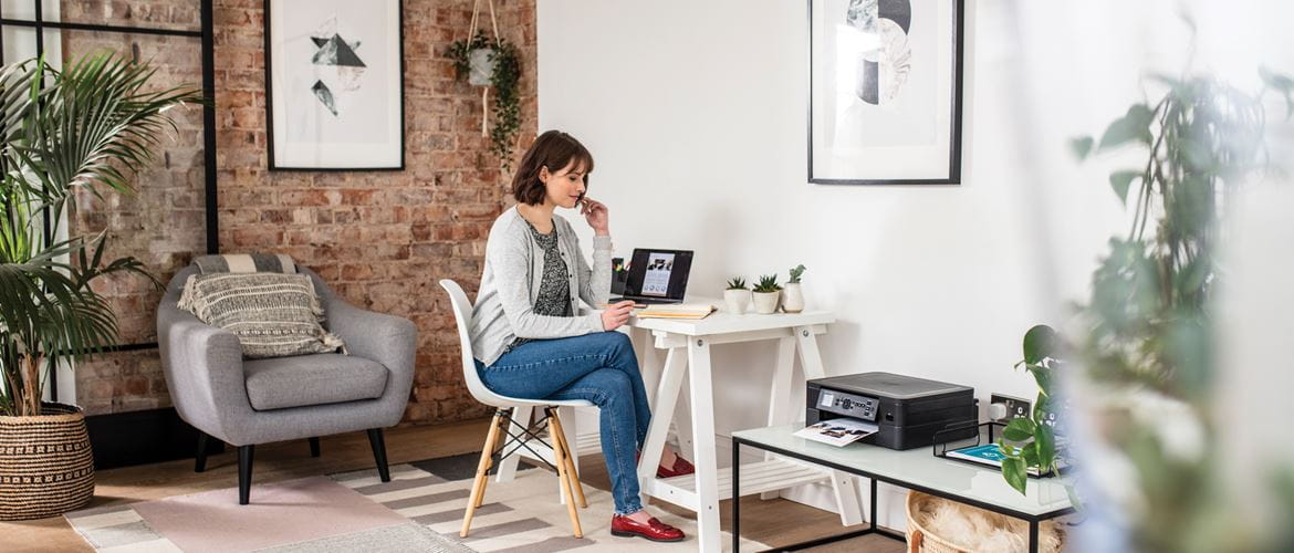 Kobieta siedzi przy biurku w domu patrząc na swój notatnik i laptopa z drukarką, obok rośliny i krzesła