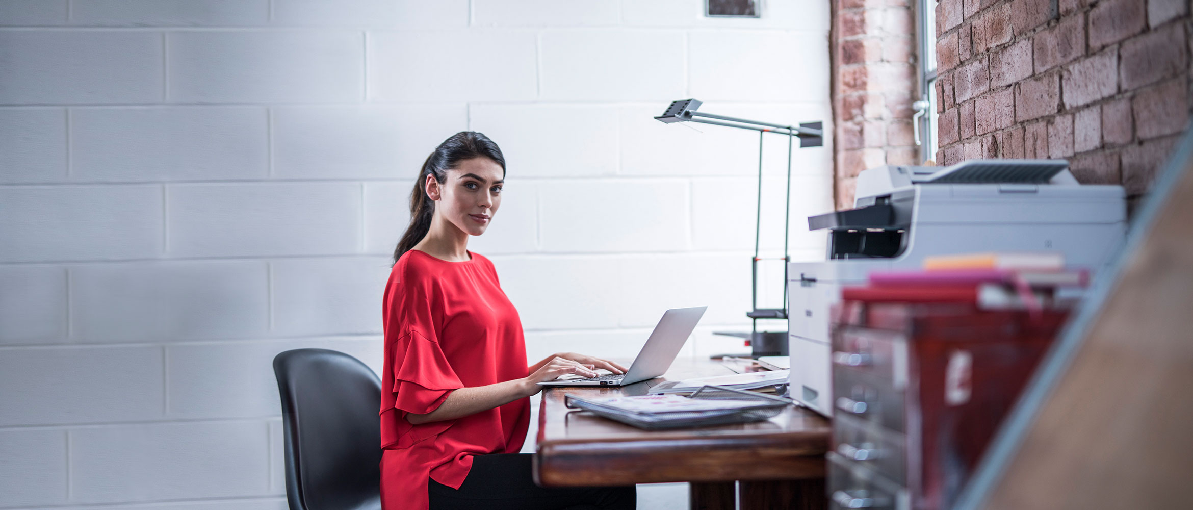 Female wearing a red top sat at a desk working on a computer around stationary with a Brother printer on the desk 