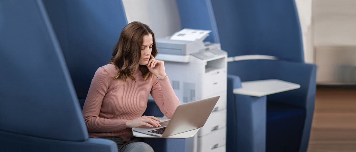 Ženska sedi v modrem naslanjaču v pisarni poleg laserske večfunkcijske naprave MFC-L6900DW
