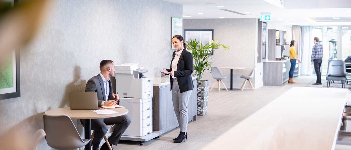 Мъж седи на маса, жена в костюм стои пред принтер в офис, столове, маси, хора на заден план 