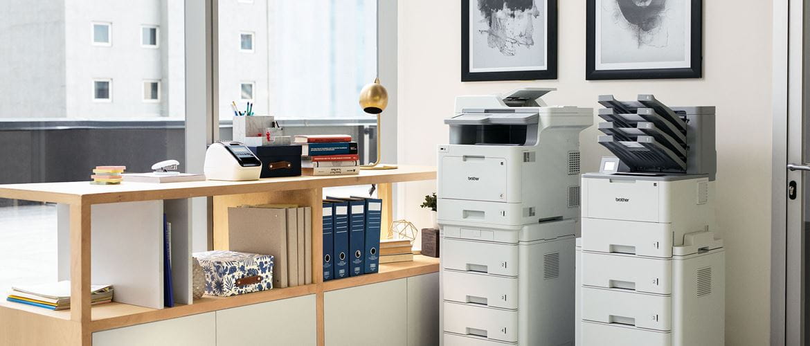 Два принтера с допълнителни тави един до друг до стена в офис, шкафове, папки, прозорци 