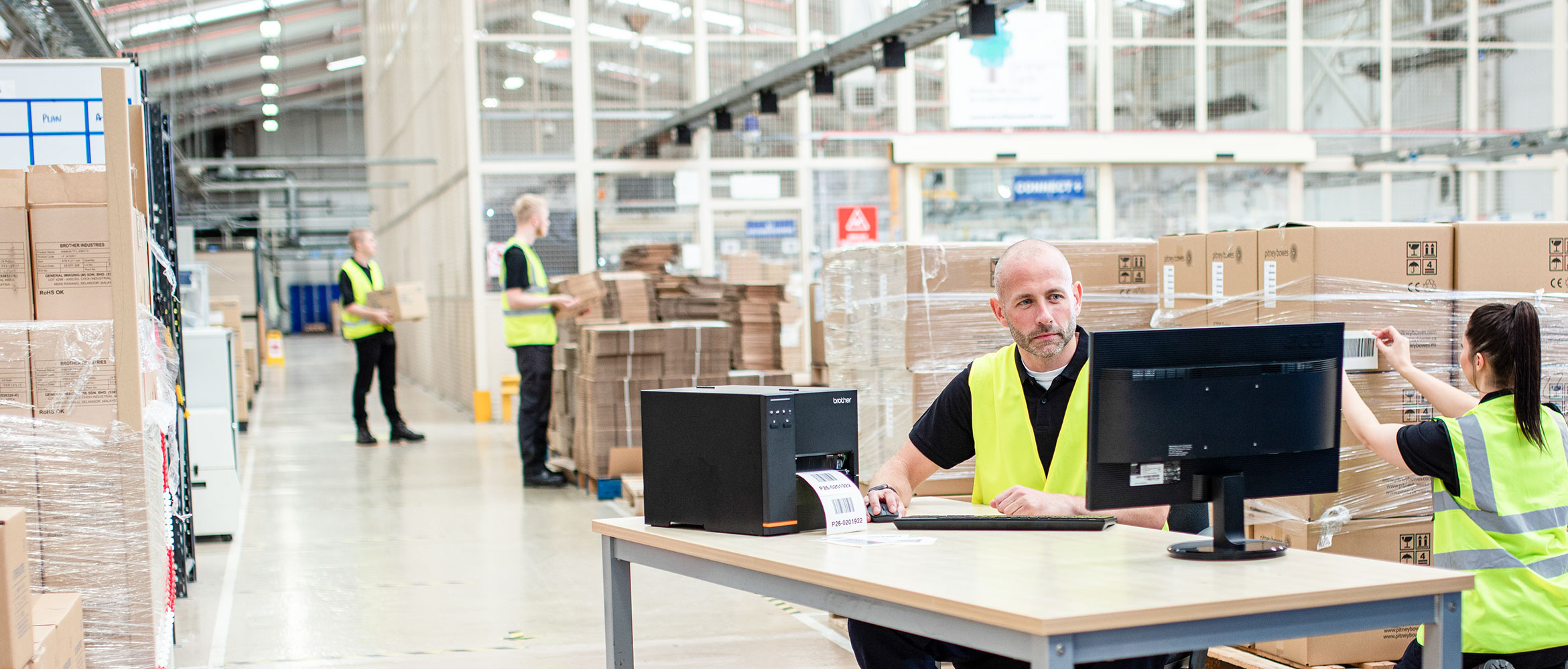 Moški v opozorilnem jopiču sedi za pisalno mizo in tiska nalepke na industrijskem tiskalniku nalepk TJ v skladišču