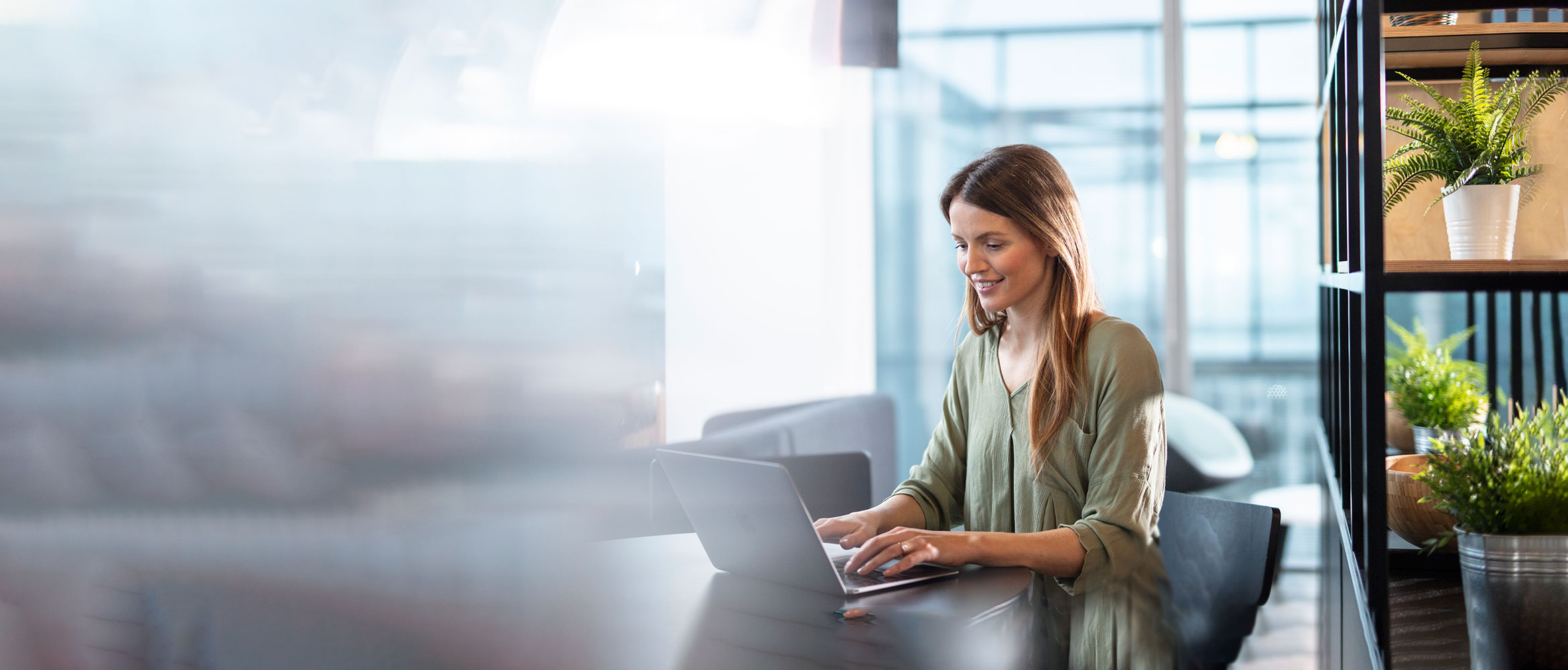 Woman sat at desk on a laptop sliming with a shelf in the background and green plants