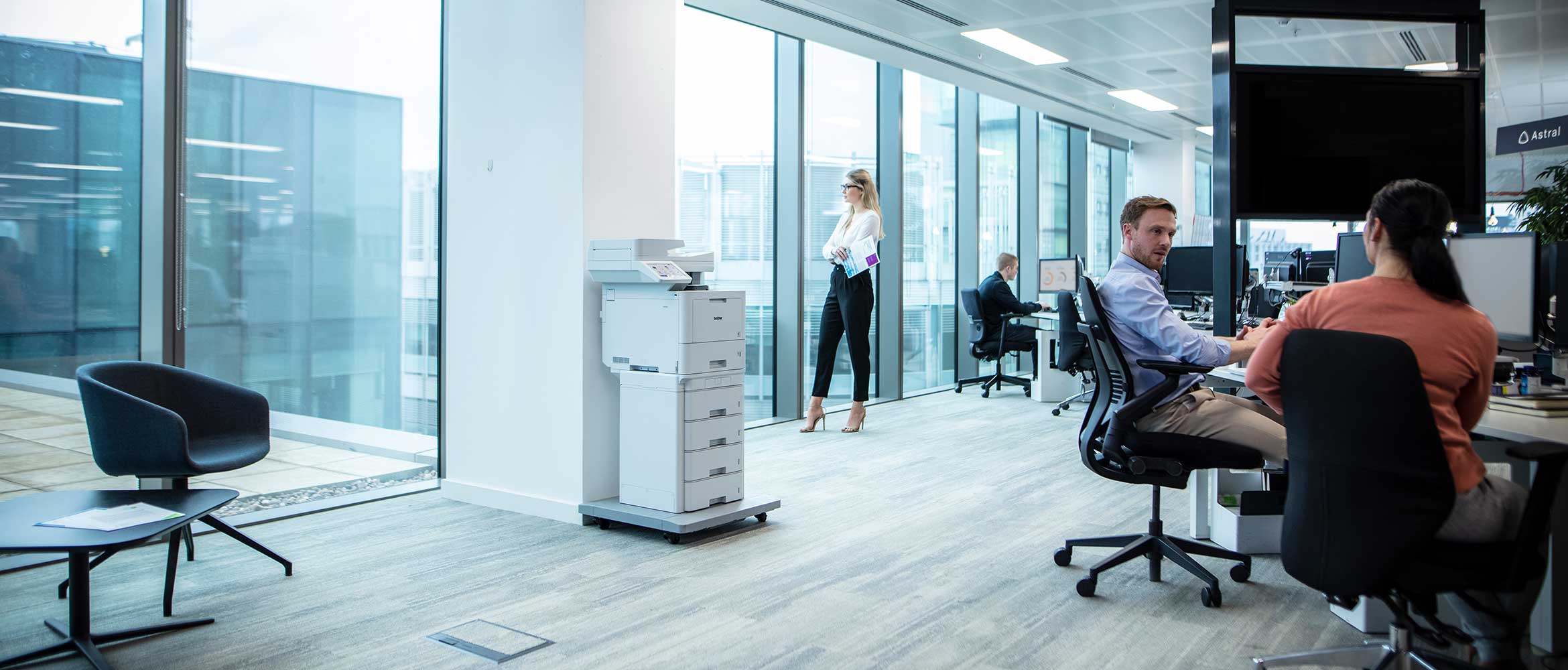 Busy office, men and women working at desks, floorstanding printer, chairs, screens, floor to ceiling windows 