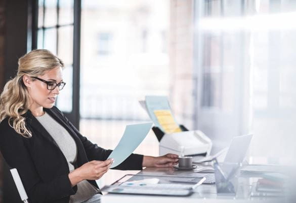 O femeie de afaceri care citește un document în timp ce stă la un birou cu un scaner lângă ea într-un mediu de birou