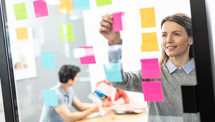 Nő színes post-it jegyzeteket helyez az üvegre, férfi és nő a háttérben, színes dokumentumok, asztal