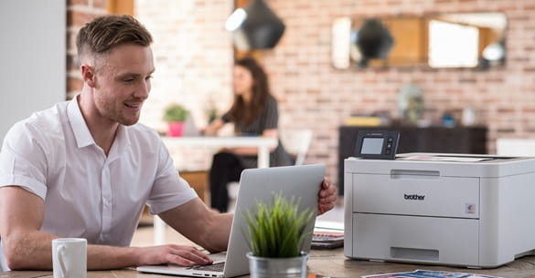 mężczyzna w białej koszulce siedzi przy stole z laptopem, obok stoi drukarka 