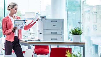 Žena odebírá dokument z multifunkční tiskárny na stole, rostliny, červené židle