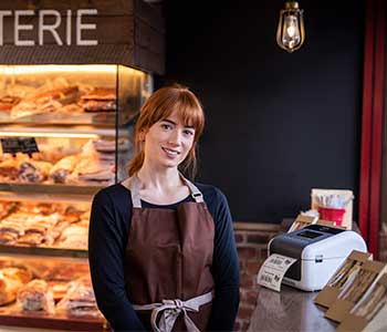 Kobieta ubrana w brązowy fartuch stoi w kawiarni, z tyłu kanapki i drukarka etykiet TD