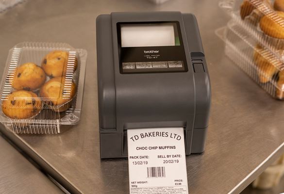 Szürke Brother címkenyomtató nyomtatott címkével a gépben, átlátszó műanyag csokoládé muffin dobozok mellett, rozsdamentes acél padon