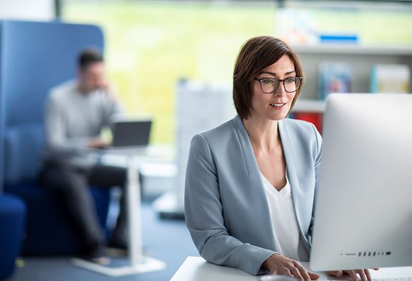 Žena s naočalama koja radi na računalu u uredu, u pozadini pisač i muškarac koji koristi laptop