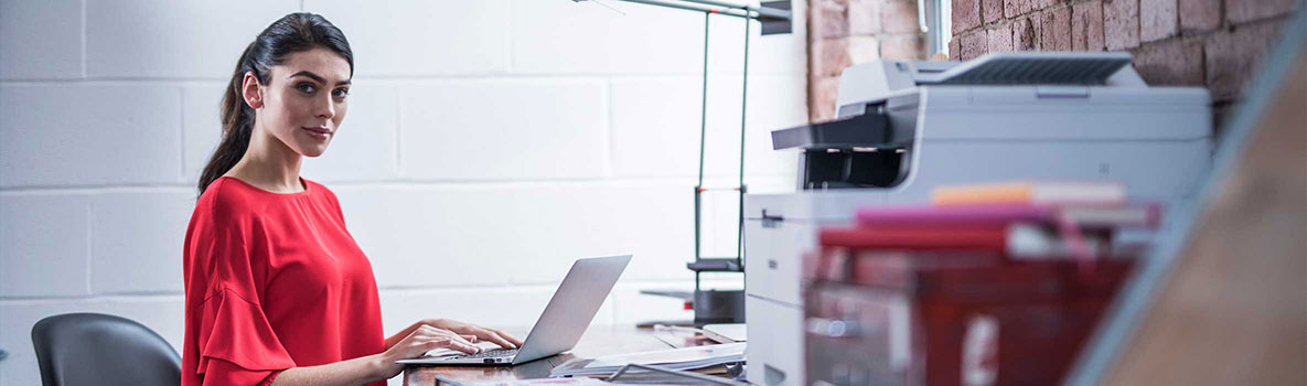 Female wearing a red top sat at a desk working on a computer around stationary with a Brother printer on the desk