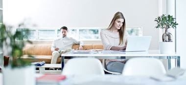 Femeie sta la birou cu laptopul, barbat sta in spate pe o canapea portocalie, plante, agende, scaune albe, fereastra