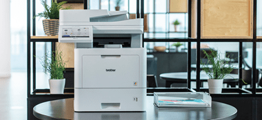 Wielofunkcyjna drukarka laserowa Brother MFC-L9670CDN stoi na okrągłym stole obok drukowanych dokumentów