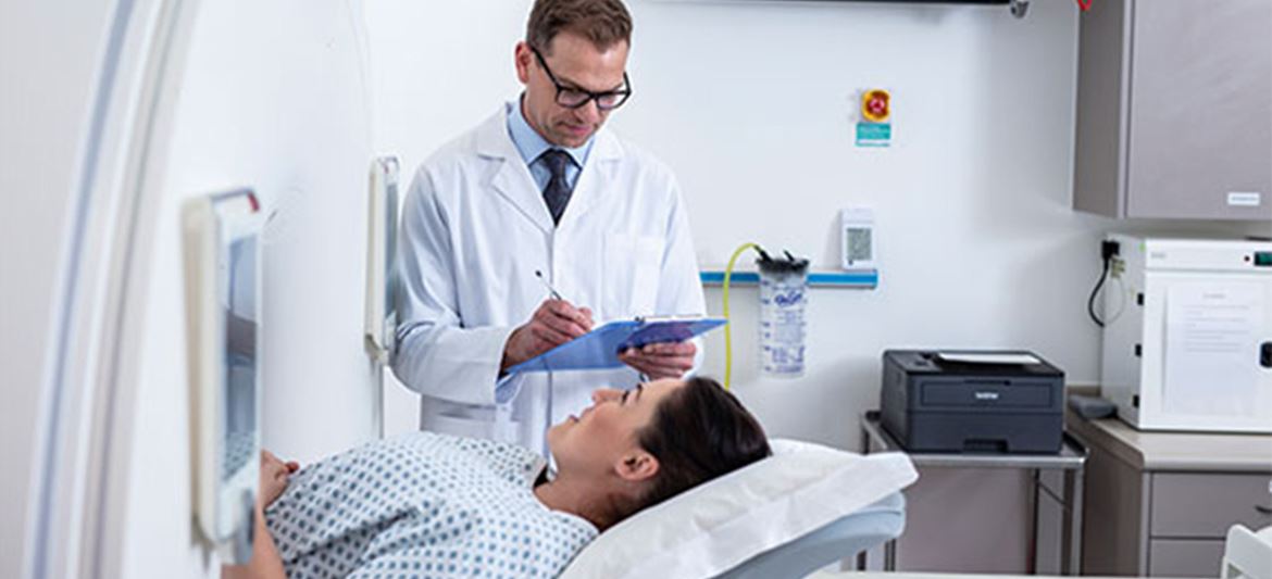 Lékař s bílým pláštěm v nemocnici u pacientky, v pozadí laserové tiskárny
