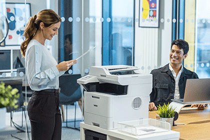 Femeie care stă lângă Brother MFC-L5710DW ținând un document mono A4 vorbind cu un bărbatul care stă la birou cu laptop, plante, scaune