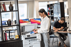 Žena v kanceláři s policí vedle multifunkční tiskárny skenuje dokument, muž v pozadí sedí u stolku