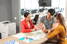 Muž a dvě ženy sedí u stolu s barevnými dokumenty v kanceláři s tiskárnou a pracovními stoly