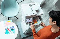 Човек натиска сензорен екран на принтер, държи цветен документ, хартия, маса, стол