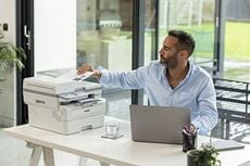 Male wearing blue shirt sat at desk using Brother MFC-L2922DW printer, laptop, plants, glass, window
