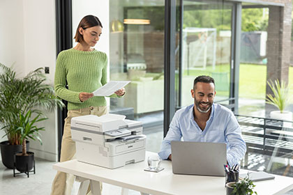 Male sat at desk using laptop next to Brother MFC-L2922DW printer, female stood holding document, plants, window