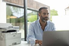 Muž sedící u stolu používající notebook, tiskárna Brother MFC-L2922DW na stole