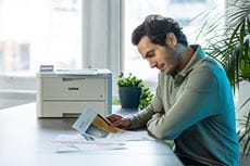 Mężczyzna siedzi przy biurku z kolorowymi dokumentami, drukarka Brother HL-L8230CDW, rośliny