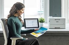 Kobieta siedzi przy biurku trzymając kolorowy dokument, laptop, drukarka Brother HL-L8240CDW