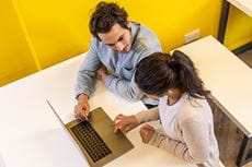 Изглед отгоре на мъж и жена, седнали един до друг с лаптоп, офис с жълта стена